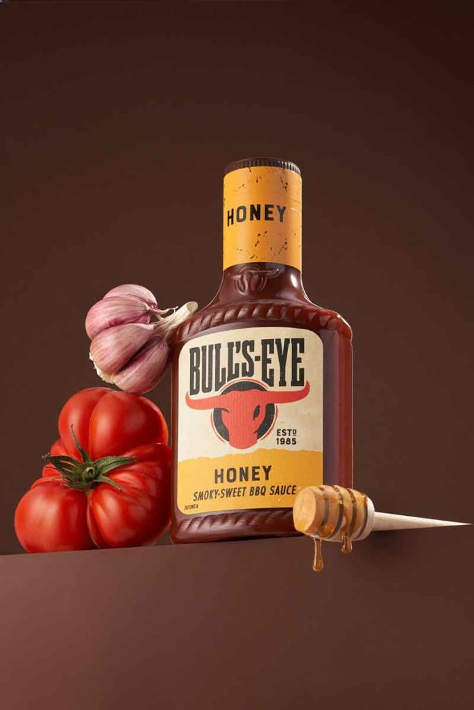 Bull’s-Eye Original BBQ Sauce bottle with garlic, red onion, and cherry tomatoes on a brown background – styled studio food image by Irina Lokteva Pikalova (Irina Lopik)