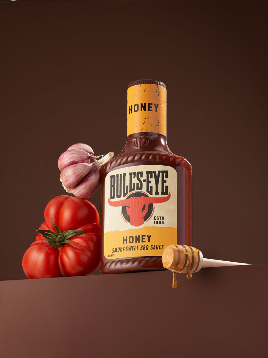 Studio shot featuring Bull’s-Eye Honey Smoky-Sweet BBQ Sauce, styled with fresh garlic, heirloom tomato, and a honey dipper. A rich brown background enhances the depth and warmth of the composition, emphasizing the product's bold and sweet character.