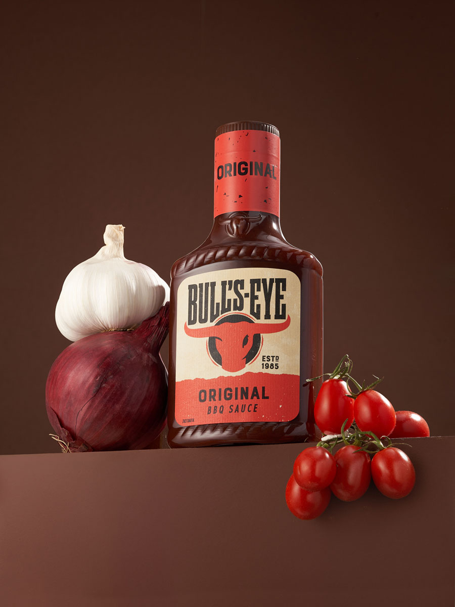 Commercial still life image showcasing Bull’s-Eye Original BBQ Sauce with garlic, red onion, and cherry tomatoes on a warm brown backdrop. The setup highlights the product’s bold flavor and classic BBQ character.